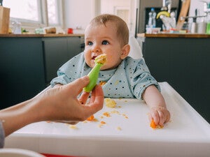 Mano de mamá dando de comer con una cuchara a su bebé que está sentado en silla alta. Alimentación complementaria del bebé.