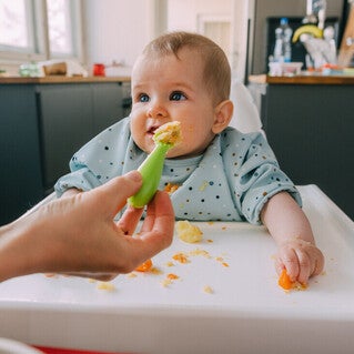 Mano de mamá dando de comer con una cuchara a su bebé que está sentado en silla alta. Alimentación complementaria del bebé.