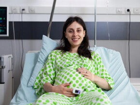 Mujer embarazada sonriendo, usando una bata de hospital y acostada en una camilla.