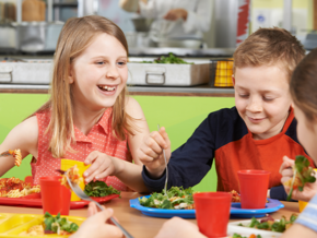 Niños comiendo en una cafetería escolar con platos de ensalada y pasta.