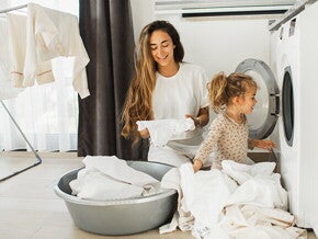 Madre e hija al lado de un lavarropas, con ropa blanca alrededor.