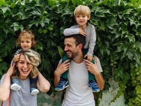 Dos adultos con dos niños sobre sus hombros, frente a una pared cubierta de hojas.