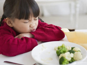 Niña pequeña sentada en la mesa en frente de un plato con brócoli y coliflor, haciendo una mueca de disgusto
