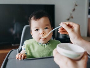 Bebé en silla alta, mano de mamá le da una cucharadita de alimento energético