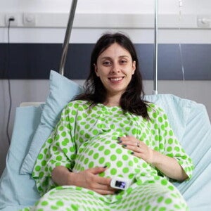 ¿Qué es la episiotomía y por qué se realiza? Mujer embarazada sonriendo, usando una bata de hospital y acostada en una camilla.