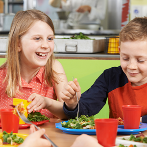 Niños comiendo en una cafetería escolar con platos de ensalada y pasta.