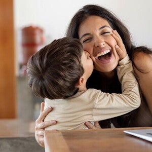 Niño dando un beso en el cachete a su madre sonriente. La mamá está sentada en frente de un ordenador portátil.