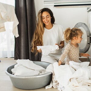 Madre e hija al lado de un lavarropas, con ropa blanca alrededor.