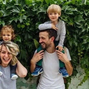 Dos adultos con dos niños sobre sus hombros, frente a una pared cubierta de hojas.