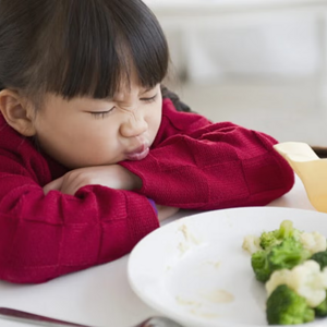 Desnutrición infantil Niña pequeña sentada en la mesa en frente de un plato con brócoli y coliflor, haciendo una mueca de disgusto