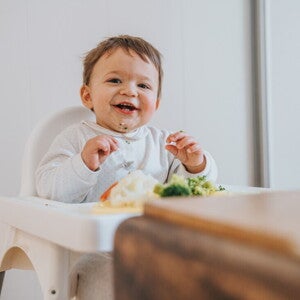 Niño de 12 meses sentado en silla alta sonriendo, con vegetales en la mesa