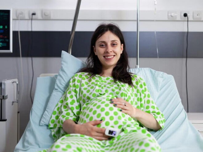 ¿Qué es la episiotomía y por qué se realiza? Mujer embarazada sonriendo, usando una bata de hospital y acostada en una camilla.