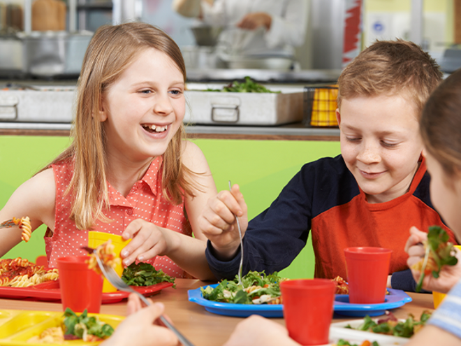 Alimentación saludable para niños escolares Niños comiendo en una cafetería escolar con platos de ensalada y pasta.