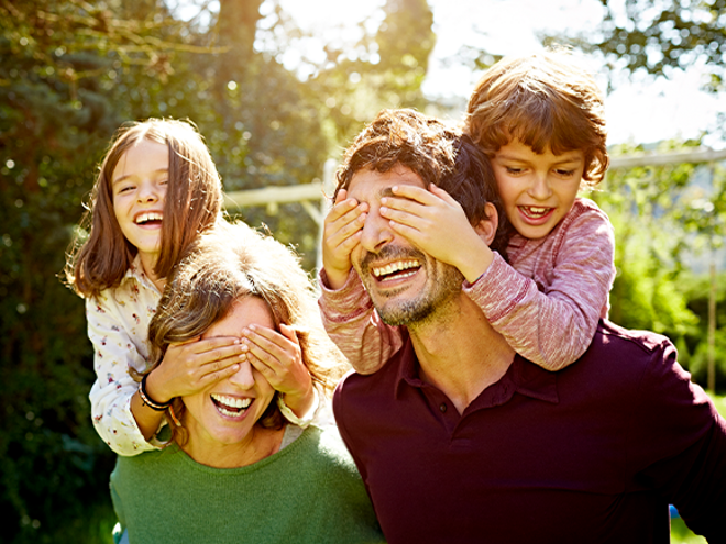 Familia de cuatro disfrutando de un día soleado al aire libre. Dos adultos llevan a dos niños sobre sus hombros, rodeados de árboles y vegetación