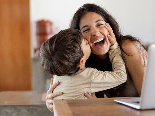 Establecer límites saludables Niño dando un beso en el cachete a su madre sonriente. La mamá está sentada en frente de un ordenador portátil.