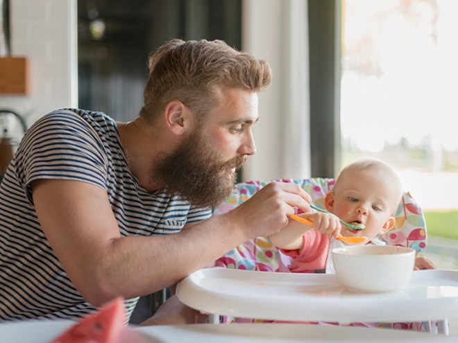 Cómo involucrar a la pareja en la alimentación del bebé Hombre con barba alimentando papilla a un bebé en una silla alta. En la mesa hay un bol blanco.