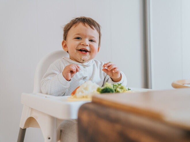 Niño de 12 meses sentado en silla alta sonriendo, con vegetales en la mesa