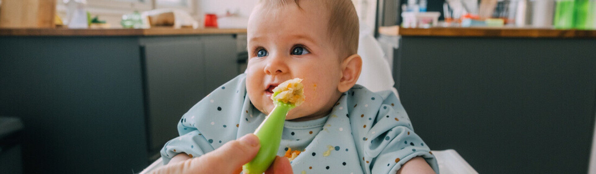 Mano de mamá dando de comer con una cuchara a su bebé que está sentado en silla alta. Alimentación complementaria del bebé.