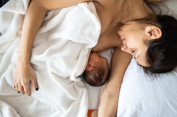 Mamá acostada en la cama, cubierta con una sábana, amamantando a su bebé recién nacido. Lactancia materna y piel con piel.